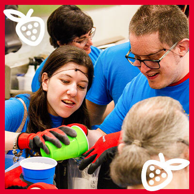 Young volunteers in blue shirts pouring a drink into cups at a community service event, smiling and helping one another.