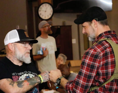 Two men in caps shake hands at a casual gathering; left man has tattoos and a black tee, right man wears a red plaid shirt.